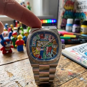 Könnte beinhalten: Eine Edelstahl-Armbanduhr mit einem farbenfrohen, künstlerischen Zifferblatt mit verschiedenen Cartoonfiguren und dem Datum 21. Die Uhr hat ein silbernes Armband und eine strukturierte Lünette. Die Uhr wird von einer Hand gehalten.
