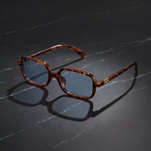 Może przedstawiać: Okulary przeciwsłoneczne w oprawie z motywem szylkretu i niebieskimi soczewkami. Prostokątne oprawki mają klasyczny wzór i są ozdobione złotymi detalami na zausznikach. Okulary leżą na ciemnej, marmurowej powierzchni.