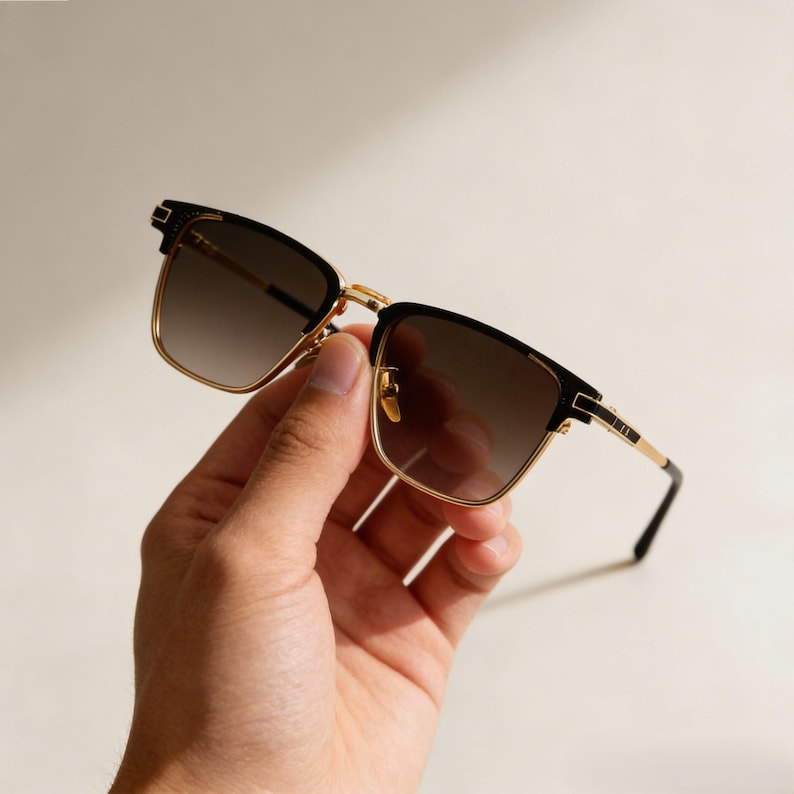 Puede incluir: Gafas de sol cuadradas con un dise&ntilde;o negro y dorado. Las gafas de sol tienen una lente degradada y se sostienen en una mano. Las monturas son negras con detalles dorados, y las lentes van de oscuro a claro.