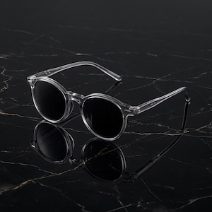 Puede incluir: Gafas de sol con montura transparente, lentes redondas y tinte oscuro. Las gafas se reflejan en una superficie oscura, similar al mármol. Las monturas y las patillas son transparentes.