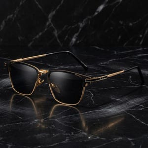 Puede incluir: Gafas de sol de montura cuadrada con montura de metal dorado y negro. Las gafas de sol tienen lentes oscuras y un dise&ntilde;o elegante y moderno. Las patillas son doradas y la montura tiene un detalle texturizado cerca de las bisagras.