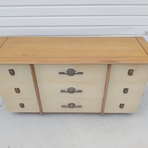 May include: A light wood dresser with six drawers and brass hardware. The dresser has a light wood top and a cream-colored body.