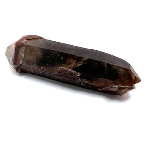 May include: A dark brown and clear quartz crystal point with a smoky brown color. The crystal is pointed at both ends and has a natural, rough texture.