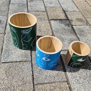 Puede incluir: Tres macetas cilíndricas de madera con diseños de caras abstractas. La maceta más grande es verde con líneas blancas, la maceta del medio es azul con líneas blancas y la maceta más pequeña es verde con líneas blancas.