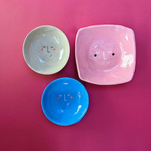 Può includere: Tre piatti in ceramica con semplici volti disegnati su di essi. I piatti sono in tonalità di rosa, blu e beige.