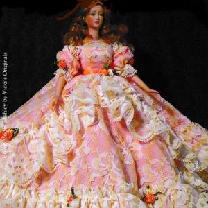 May include: A doll wearing a pink and white lace dress with a full skirt and puffed sleeves. The dress is decorated with small orange roses and a peach-coloured sash. The doll has red hair and is labelled "Anne Marie" and "A Pattern to Fit 25.4 cm Coquette Cissy."