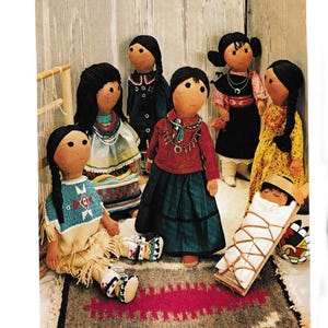 May include: A collection of handmade Native American dolls, each with unique clothing and hairstyles. The dolls are dressed in traditional attire, including beaded tops, skirts, and dresses. One doll is cradling a baby doll in a wooden cradle. The dolls are arranged on a patterned rug.