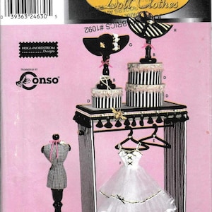 May include: Simplicity Series 4 Couturier Doll Clothes sewing pattern. The image shows a doll dress, a dress form, and a display stand. The stand has black and white striped fabric and a black trim. The dress is white with black accents.