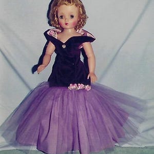 May include: A vintage doll wearing a purple velvet dress with a tulle skirt and a pink flower accent. The doll has blonde hair and blue eyes.