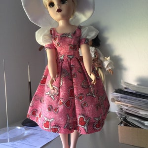 Puede incluir: Una muñeca de porcelana con un vestido rosa con estampado de corazones y un sombrero blanco. La muñeca tiene el pelo rubio y los ojos azules.