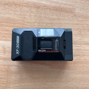 Puede incluir: Una cámara de película Ricoh XF-30 negra con una lente Ricoh Rikenon 1:1.4 f=35mm. La cámara tiene un flash incorporado y un dial de velocidad de película que llega hasta 1000 ISO.