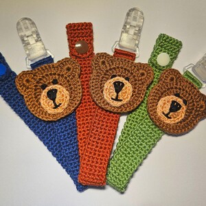 May include: Four crocheted bear-shaped teethers with different colored straps. The teethers are brown and have black eyes and noses. The straps are blue, orange, green, and white.