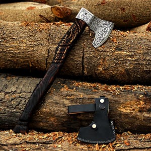 May include: A black-handled axe with a silver blade featuring intricate knotwork designs. The axe is resting on a pile of logs and comes with a black leather sheath.