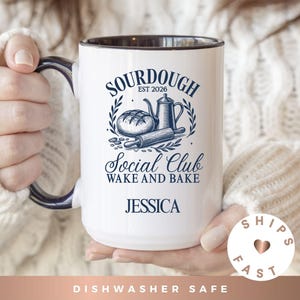 May include: White ceramic mug with a black rim and handle, featuring a blue design with the words "SOURDOUGH EST 2026" and "Social Club WAKE AND BAKE". The name "JESSICA" is printed below the design. The mug is dishwasher safe.
