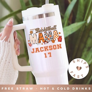 May include: White insulated tumbler with a handle, featuring the text "Basketball MAMA JACKSON 17" in orange and brown. The design includes basketball-themed graphics. The tumbler has a white lid and handle.