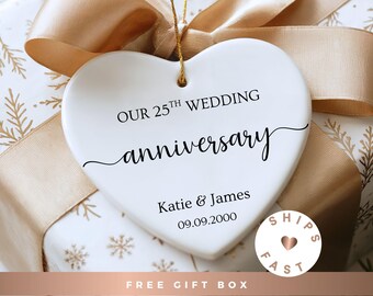 May include: A white heart-shaped ornament with the text "OUR 25TH WEDDING anniversary" in black script. Below, it reads "Katie & James 09.09.2000." The ornament hangs from a gold string, set against a gift box with a gold ribbon.
