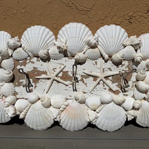 May include: A decorative wall-mounted coat rack featuring a distressed white wooden base, adorned with various white seashells and starfish. It includes four metal hooks for hanging items. The rack is designed with a coastal theme.
