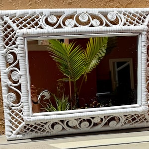 Puede incluir: Espejo rectangular blanco con marco de mimbre tejido. El marco presenta patrones decorativos de volutas y enrejado. El espejo refleja una escena tropical con hojas de palmera verdes y una pared roja.