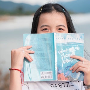 May include: A young woman is holding a blue book open in front of her face. The book cover has the title "Minh nghi gi khi hanh phuc!" and the author "Rosie Nguyen".