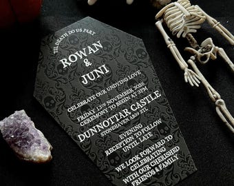 May include: Black and white wedding invitation with a coffin-shaped design. The invitation features a gothic font and skull imagery. The text reads "Til Death Do Us Part, Rowan & Juni, Celebrate Our Undying Love, Friday 13th November 2026, Ceremony to Begin at 3PM, Dunnottar Castle, Stonehaven AB39 2TL, Evening Reception to Follow Until Late, We Look Forward to Celebrating with Our Cherished Friends & Family."