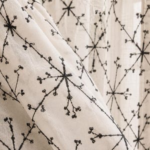 Puede incluir: Primer plano de una cortina blanca con un patrón floral bordado en negro. El diseño presenta explosiones de estrellas y delicadas ramas con pequeños capullos. La tela es ligeramente texturizada y la cortina está parcialmente drapeada.