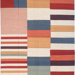 May include: A rectangular rug with a patchwork design. The rug features blocks of solid colours like red, blue, and beige, and sections with horizontal stripes in various colours. The overall design is a mix of geometric shapes and patterns.