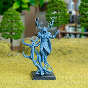 May include: A blue miniature figure of a hooded character with antlers, standing on a black base. The figure is holding a cloth and is surrounded by miniature trees and a green surface. The figure is a fantasy character.