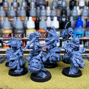 May include: A collection of gray miniature fantasy figures, each mounted on a stylized creature. The figures are on black bases and appear to be unpainted. The background includes various paint bottles, suggesting a hobby or gaming theme.