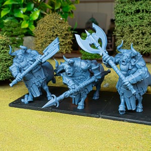 May include: Three blue fantasy figurines of bull-headed warriors with weapons. Each figure has horns, armor, and is holding a large axe or spear. They are standing on a black base, with green foliage in the background.