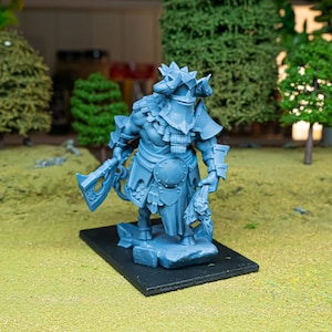 May include: A light blue miniature figurine of a warrior in armor, holding an axe and a severed head. The figurine is on a black base, set against a backdrop of miniature trees and grass.