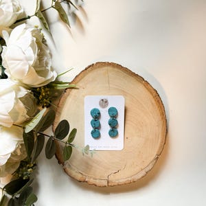 May include: Turquoise and black marbled dangle earrings displayed on a white card, resting on a wooden slice. The earrings have three stacked circular elements. White flowers and greenery are arranged around the wooden slice.