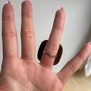 K&ouml;nnte beinhalten: Ein silberner Ring mit einem gro&szlig;en, dunkelbraunen, rechteckigen Stein. Der Ring wird an einer Hand mit rosa Nagellack getragen.