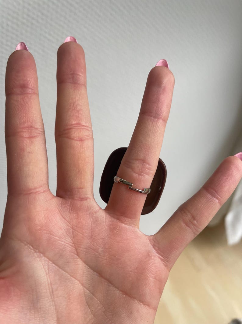 K&ouml;nnte beinhalten: Ein silberner Ring mit einem gro&szlig;en, dunkelbraunen, rechteckigen Stein. Der Ring wird an einer Hand mit rosa Nagellack getragen.