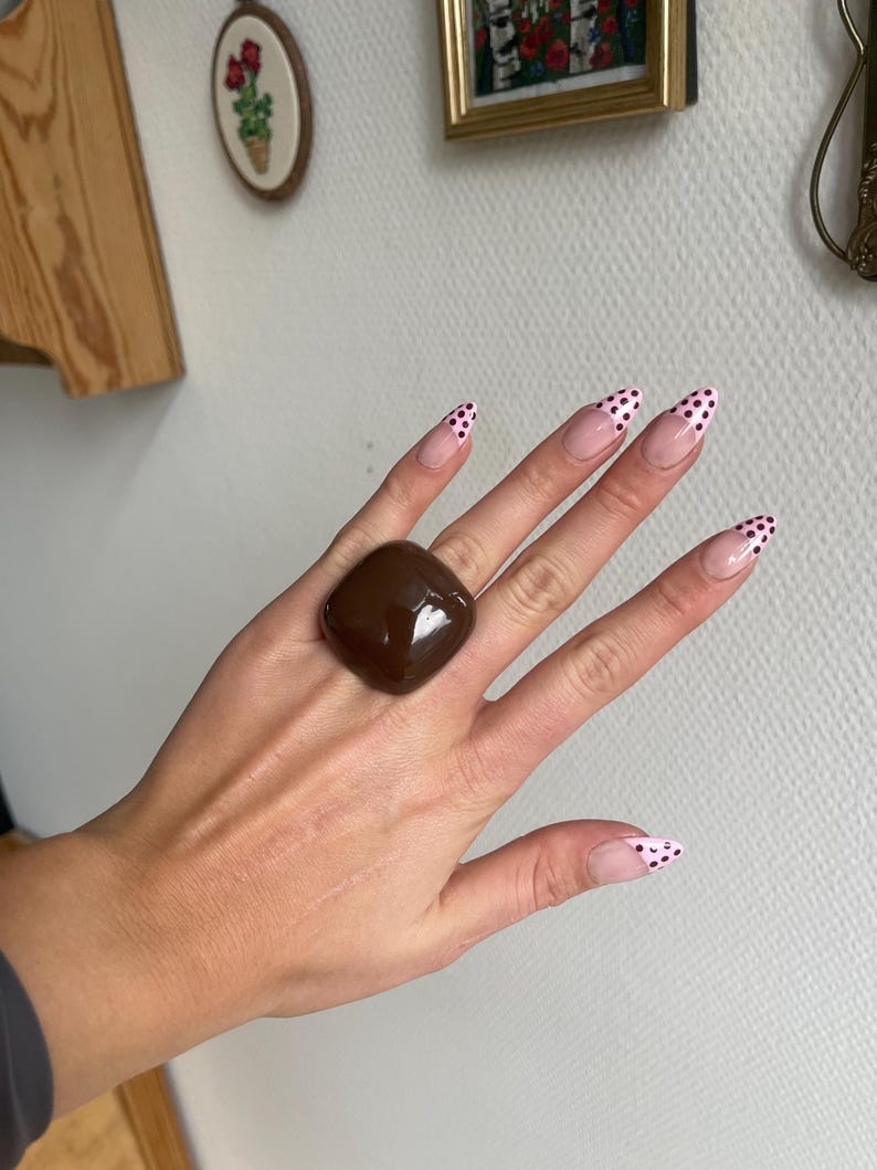K&ouml;nnte beinhalten: Ein gro&szlig;er, quadratischer, dunkelbrauner Ring mit gl&auml;nzendem Finish. Der Ring ist der Blickfang, pr&auml;sentiert auf einer Hand mit langen, spitzen N&auml;geln, die mit rosa und schwarzen Punkten verziert sind. Der Hintergrund umfasst Wanddekorationen.