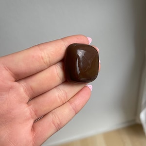K&ouml;nnte beinhalten: Ein polierter, dunkelbrauner Edelstein mit abgerundeten Kanten. Der Stein hat eine glatte, reflektierende Oberfl&auml;che und wird in einer Hand gehalten. Die Farbe des Edelsteins ist ein sattes, tiefes Braun, und er scheint ein Naturstein zu sein.