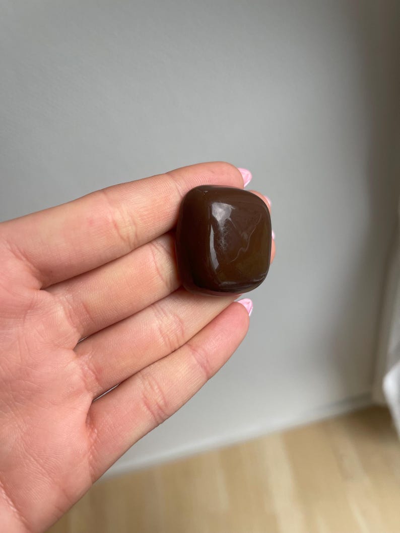 K&ouml;nnte beinhalten: Ein polierter, dunkelbrauner Edelstein mit abgerundeten Kanten. Der Stein hat eine glatte, reflektierende Oberfl&auml;che und wird in einer Hand gehalten. Die Farbe des Edelsteins ist ein sattes, tiefes Braun, und er scheint ein Naturstein zu sein.