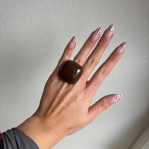 K&ouml;nnte beinhalten: Eine Hand tr&auml;gt einen gro&szlig;en, quadratischen, braunen Ring. Die langen, mandelf&ouml;rmigen N&auml;gel sind mit einer hellrosa Basis und schwarzen Tupfen auf den Spitzen bemalt. Der Ring hat eine glatte, polierte Oberfl&auml;che und ist der Fokus.