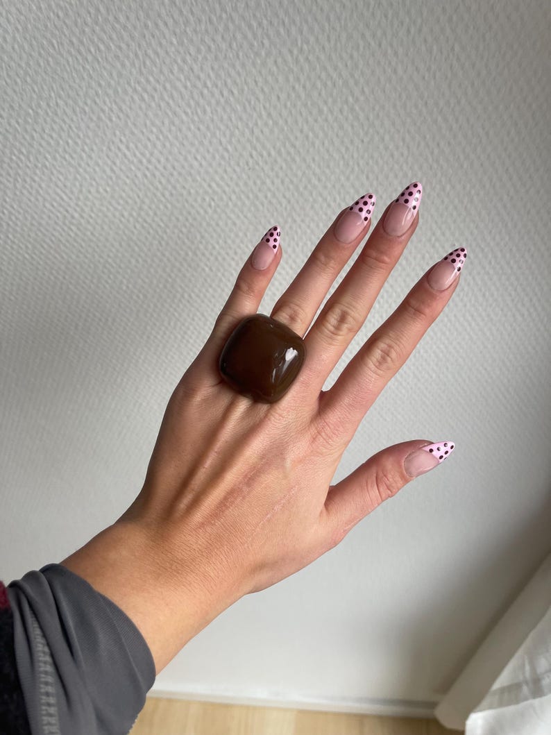 K&ouml;nnte beinhalten: Eine Hand tr&auml;gt einen gro&szlig;en, quadratischen, braunen Ring. Die langen, mandelf&ouml;rmigen N&auml;gel sind mit einer hellrosa Basis und schwarzen Tupfen auf den Spitzen bemalt. Der Ring hat eine glatte, polierte Oberfl&auml;che und ist der Fokus.