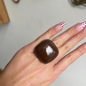 K&ouml;nnte beinhalten: Ein gro&szlig;er, quadratischer, dunkelbrauner Ring mit gl&auml;nzendem Finish. Der Ring ist der Blickfang, vor einem neutralen Hintergrund. Die Form des Rings ist geometrisch und k&uuml;hn, was ihn zu einem auff&auml;lligen Schmuckst&uuml;ck macht.