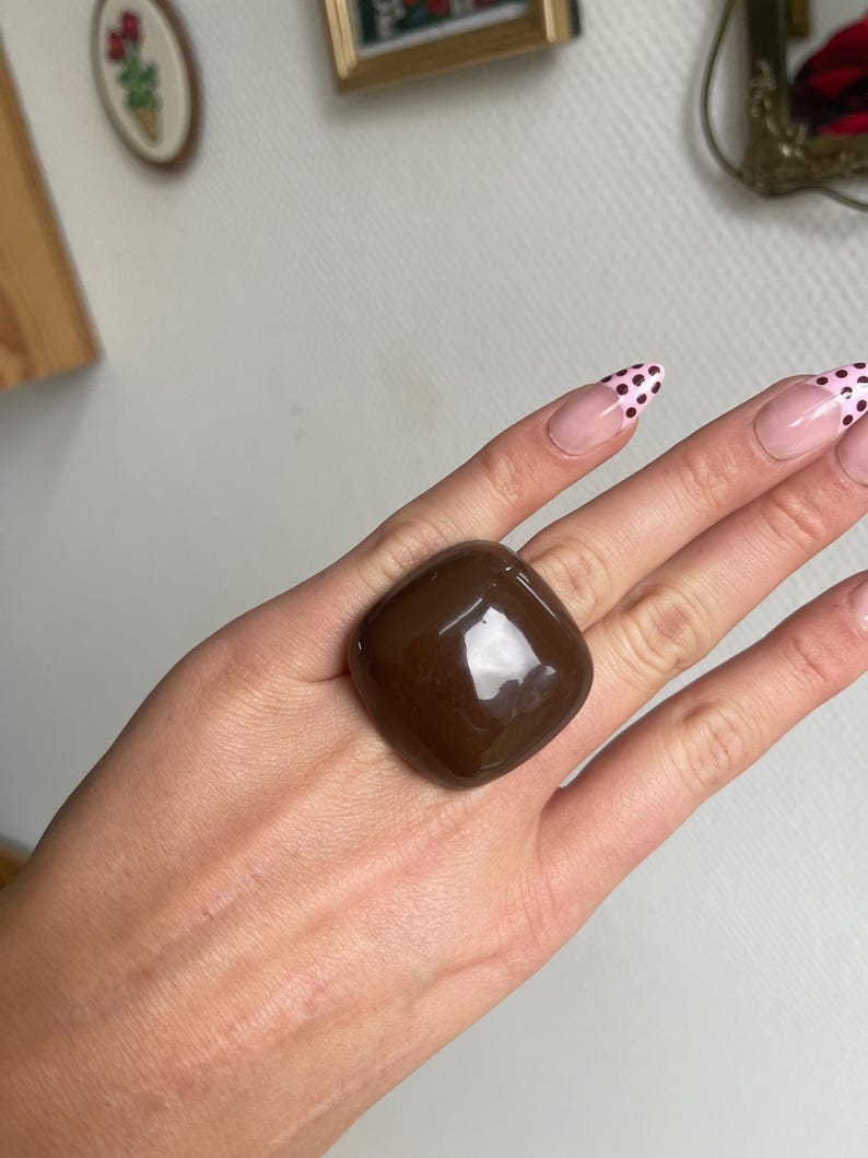 K&ouml;nnte beinhalten: Ein gro&szlig;er, quadratischer, dunkelbrauner Ring mit gl&auml;nzendem Finish. Der Ring ist der Blickfang, vor einem neutralen Hintergrund. Die Form des Rings ist geometrisch und k&uuml;hn, was ihn zu einem auff&auml;lligen Schmuckst&uuml;ck macht.