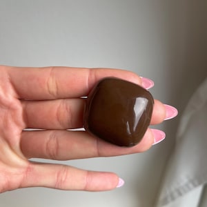 K&ouml;nnte beinhalten: Ein glatter, dunkelbrauner, rechteckiger Stein mit abgerundeten Kanten. Der Stein hat eine gl&auml;nzende Oberfl&auml;che, die das Licht reflektiert. Das Objekt wird in einer Hand mit rosa lackierten N&auml;geln gehalten.