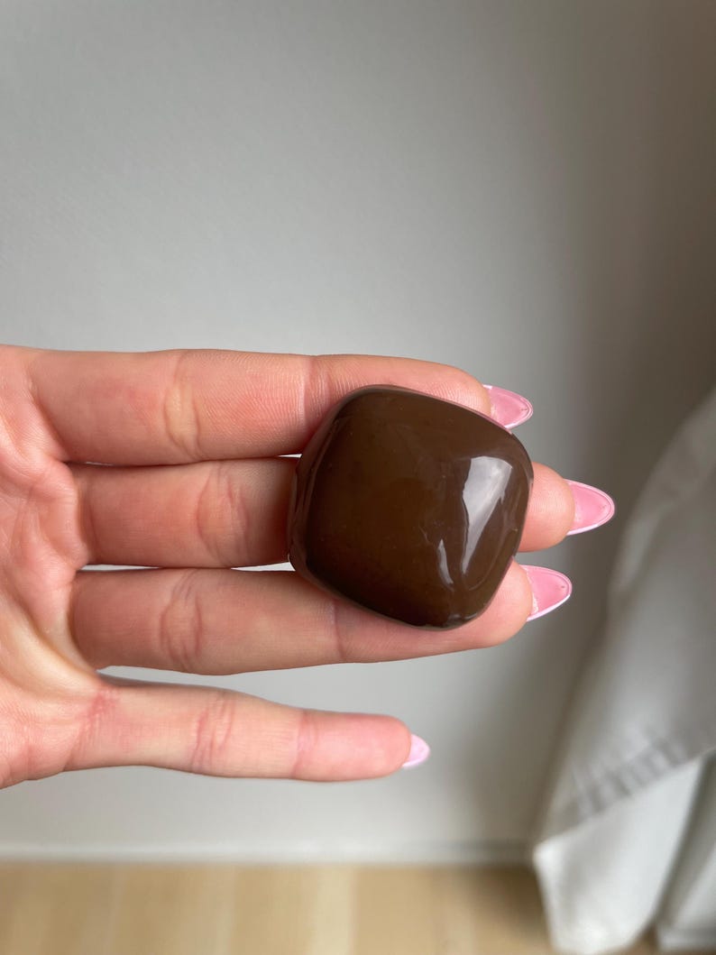 K&ouml;nnte beinhalten: Ein glatter, dunkelbrauner, rechteckiger Stein mit abgerundeten Kanten. Der Stein hat eine gl&auml;nzende Oberfl&auml;che, die das Licht reflektiert. Das Objekt wird in einer Hand mit rosa lackierten N&auml;geln gehalten.