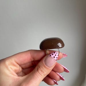 K&ouml;nnte beinhalten: Ein brauner, pilzf&ouml;rmiger Ring mit gl&auml;nzendem Finish. Der Ring hat ein d&uuml;nnes, goldfarbenes Band. Der Pilzaufsatz ist einfarbig dunkelbraun. Der Ring ist ein einzigartiges Schmuckst&uuml;ck.