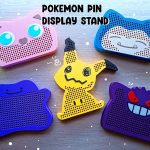 Puede incluir: Cinco coloridos expositores de pines de Pokémon. Los expositores tienen forma de personajes de Pokémon, como Jigglypuff, Snorlax, Mimikyu, Ditto y Gengar. Los expositores son rosas, azules, amarillos, morados y azules.