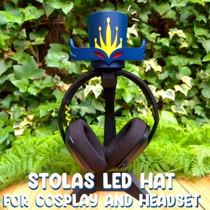 Puede incluir: Un soporte de auriculares negro con un sombrero LED azul y amarillo en la parte superior. El sombrero tiene una corona y un diseño de máscara. El texto "Stolas LED Hat for Cosplay and Headset" está impreso debajo del soporte.
