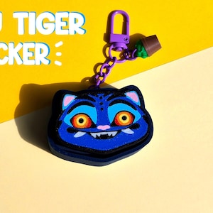May include: A blue tiger-faced keyring with orange eyes and a purple clasp. The keyring has the text "DERPY TIGER CLICKER" in white. The background is yellow and cream.