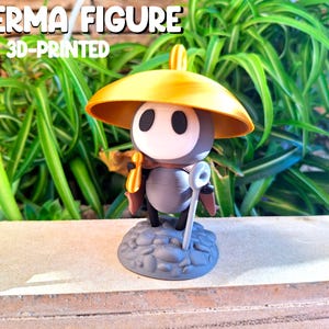 Può includere: Figura di Sherma stampata in 3D, con cappello giallo e bastone. La figura ha un viso bianco con occhi neri e si trova su una base di pietra grigia. Lo sfondo presenta piante verdi. Il testo "SHERMA FIGURE 3D-PRINTED" è visibile.