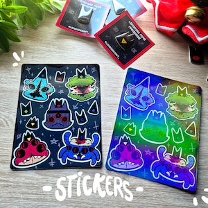 Puede incluir: Dos hojas de pegatinas holográficas con criaturas de dibujos animados coloridas con múltiples ojos. Las pegatinas están sobre una superficie de madera con el texto "STICKERS" impreso debajo.