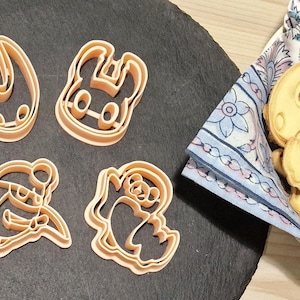 Puede incluir: Cinco cortadores de galletas con forma de personajes del videojuego Hollow Knight. Los cortadores están hechos de plástico marrón claro y están dispuestos sobre una superficie negra.
