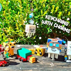 Op de afbeelding: Een verzameling gepixelde karakter sleutelhangers met bedels. De sleutelhangers zijn in verschillende kleuren, waaronder rood, geel, groen en wit. De afbeelding bevat de tekst "KEYCHAINS WITH CHARMS" en "IT COMES WITH THIS TYPE OF CHAIN".